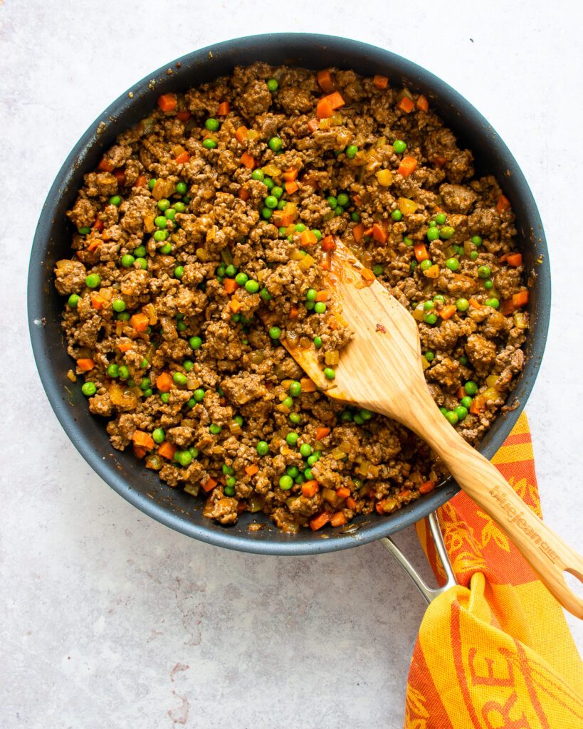 Shepherd's pie filling in a skillet with a wooden spoon and a towel wrapped around the handle.