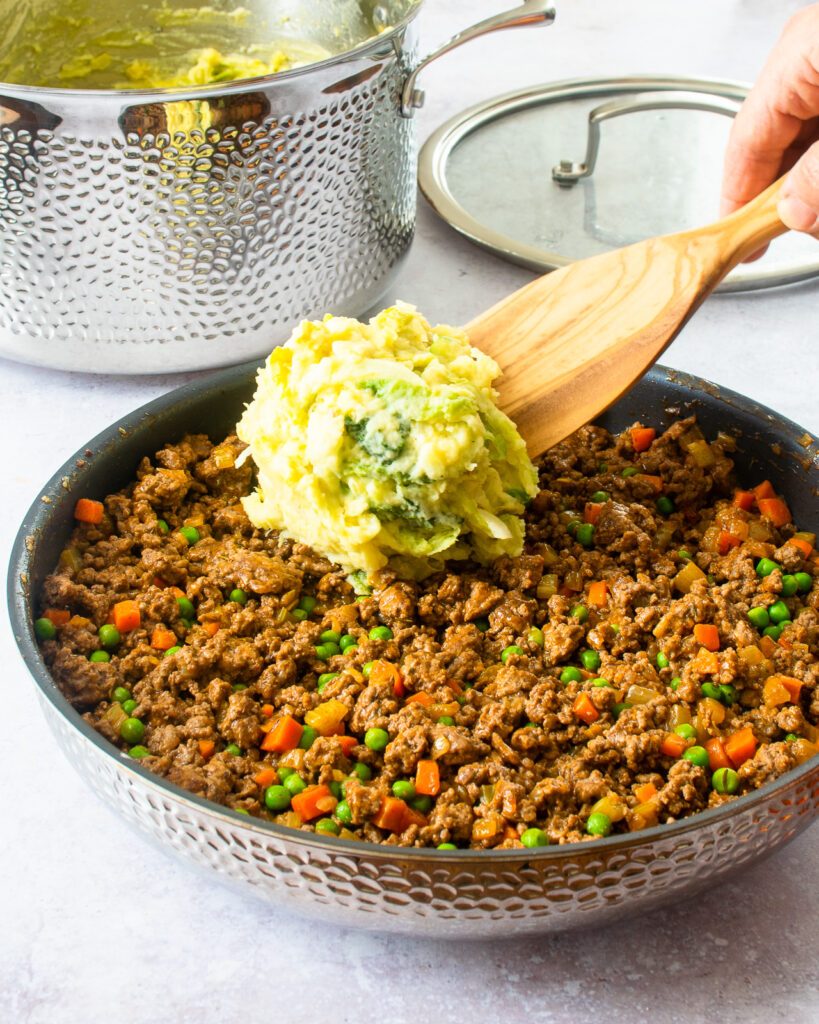 Scooping mashed potatoes on top of shepherd's pie filling in a stainless steel skillet with a stainless steel stockpot in the background.