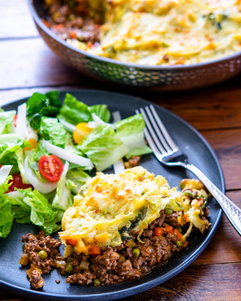 Shepherd's pie on a black plate with salad and a fork and a skillet of more shepherd's pie in the background.