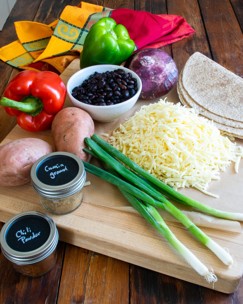 Ingredients on a cutting board on a wooden table - red onion, bell peppers, black beans, sweet potatoes, spices, green onions, grated cheese and tortillas.