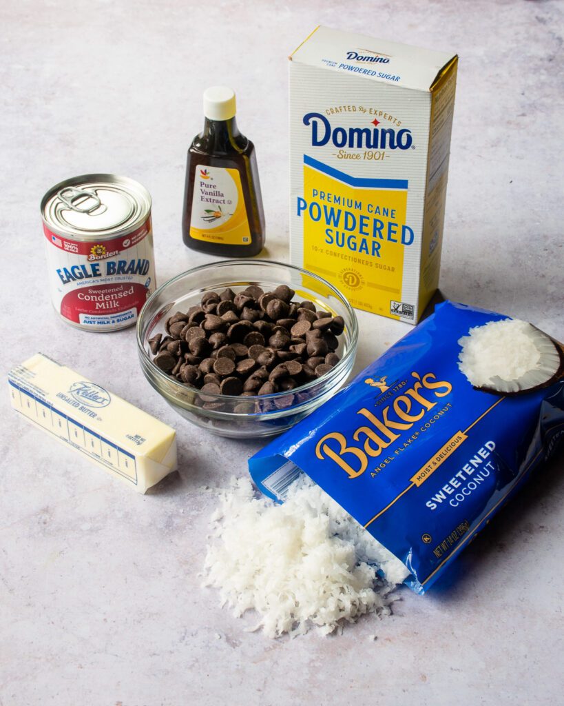Ingredients on a countertop - sweetened coconut, chocolate chips, butter, condensed milk, vanilla extract and powdered sugar.