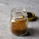 A jar of spices with a lid in the background on a gray counter.