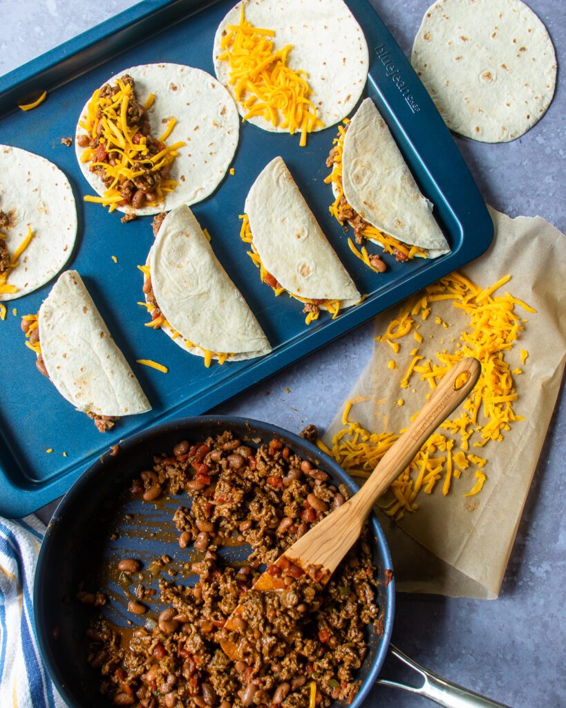 Looking down on tacos being assembled - a skillet of taco mix, some grated cheese and tortillas in various stages of being filled.