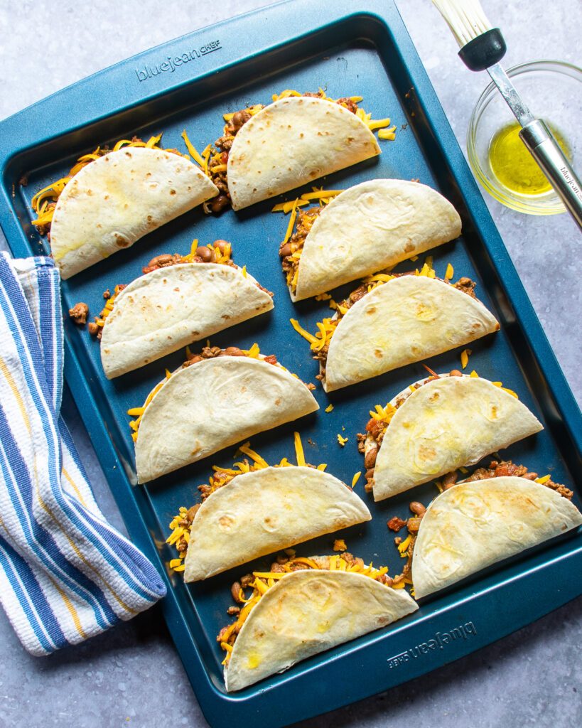 Assembled tacos on a blue sheet pan, ready for frying.