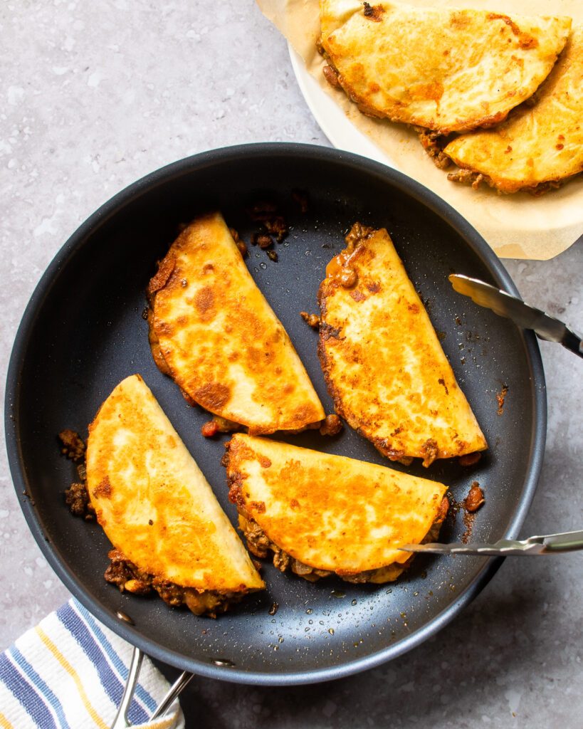Crispy beef tacos frying in a large skillet with tongs resting on the side.