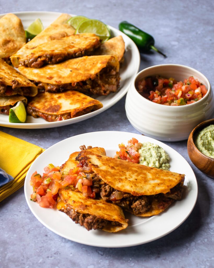 A white plate with two crispy beef tacos, salsa and guacamole with more tacos on a platter in the background.