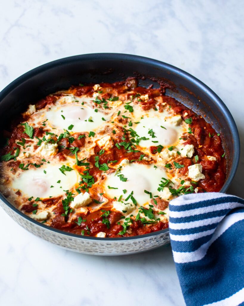 Shakshuka in a hammered stainless steel skillet with a blue and white striped towel around the handle.