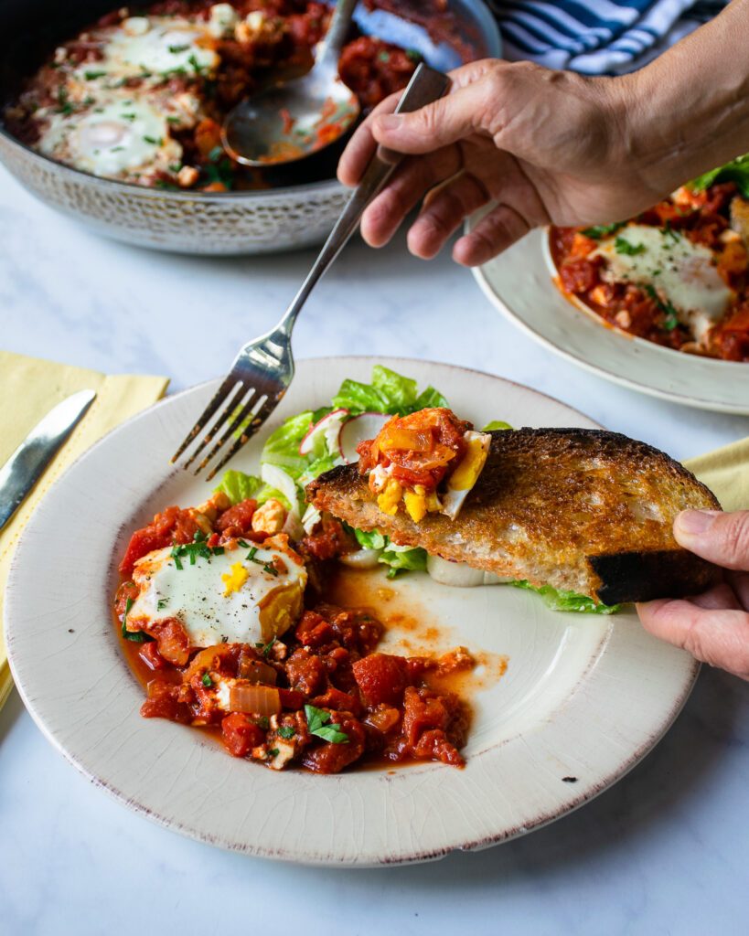 Two hands eating shakshuka off a plate with a piece of toast and a fork.