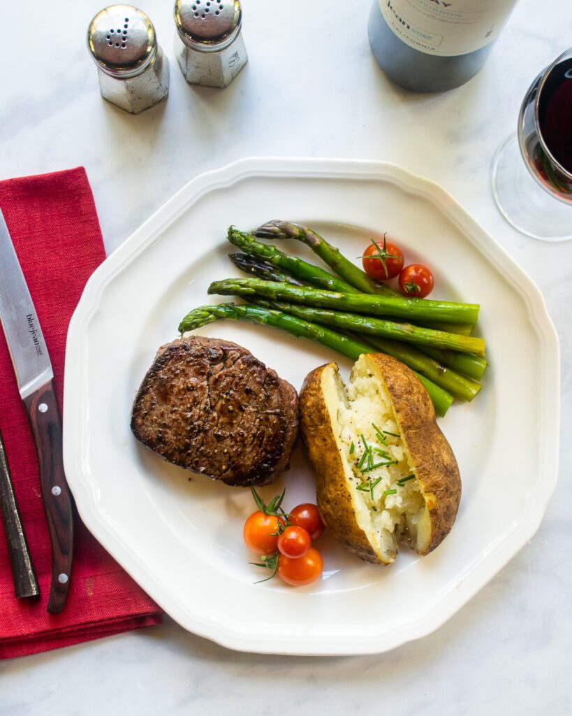 Looking down on a white plate with filet mignon, baked potato and asparagus, place setting and wine.