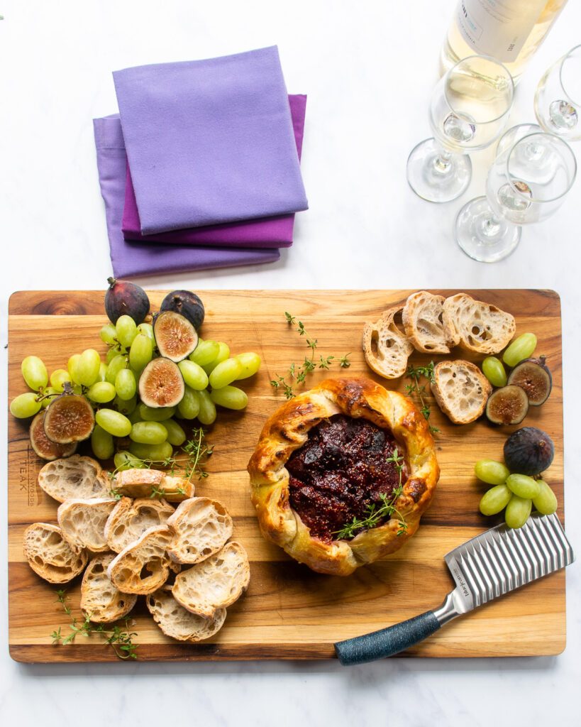 Baked Brie with Fig jam on a serving board with crostini, grapes, raw figs and a cheese knife, with wine and napkins next to it.