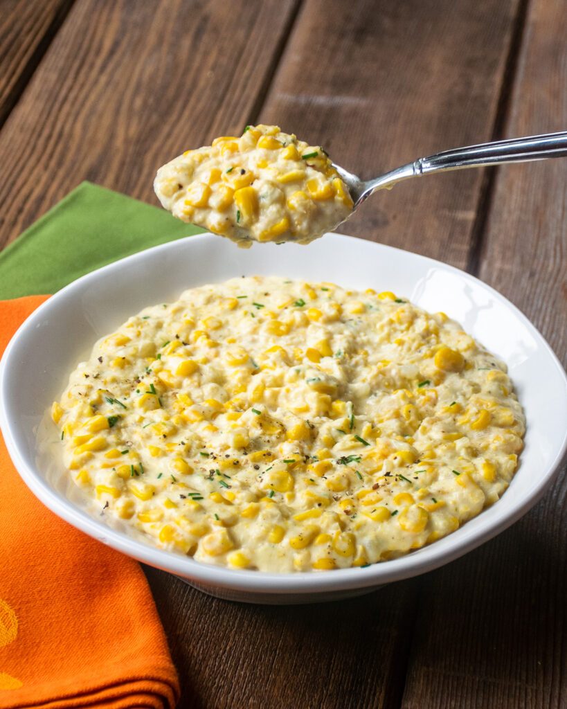 A spoon lifting creamy corn out of a white bowl with orange and green napkins next to it.