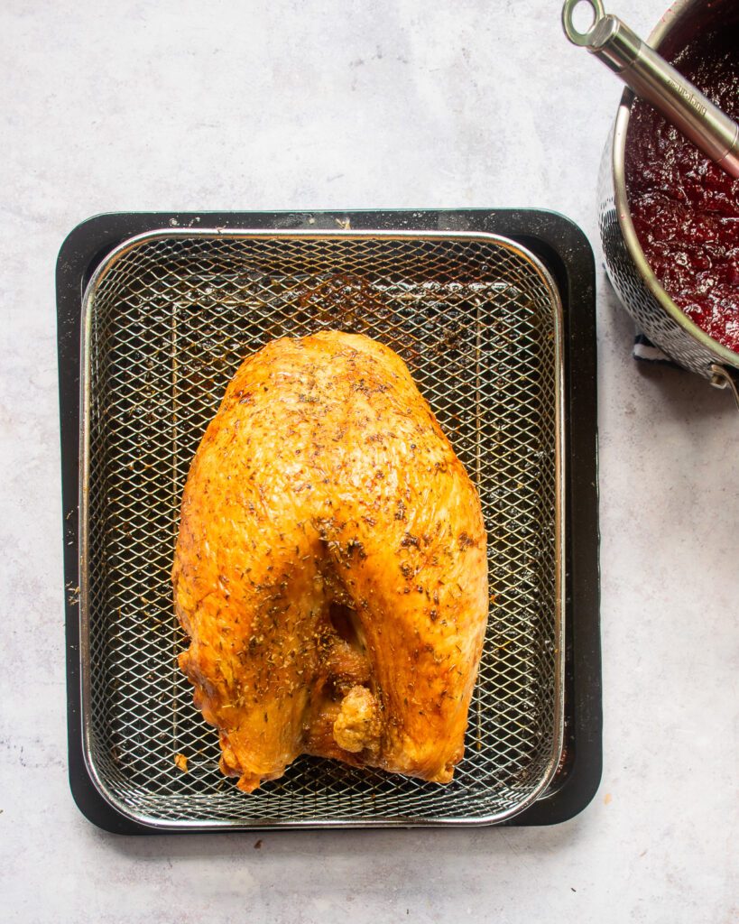 Looking down on a roast turkey breast in an air fryer basket with a saucepan of cranberry sauce on the side.