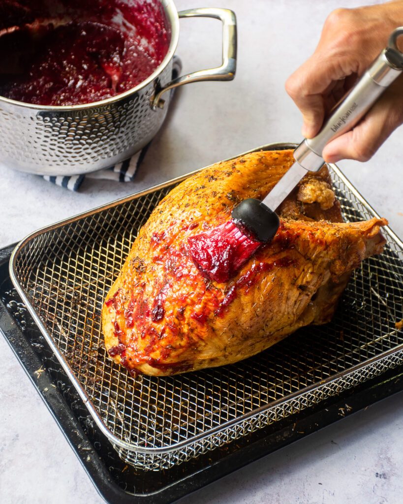 A hand brushing cranberry glaze on a roast turkey breast in an air fryer basket.