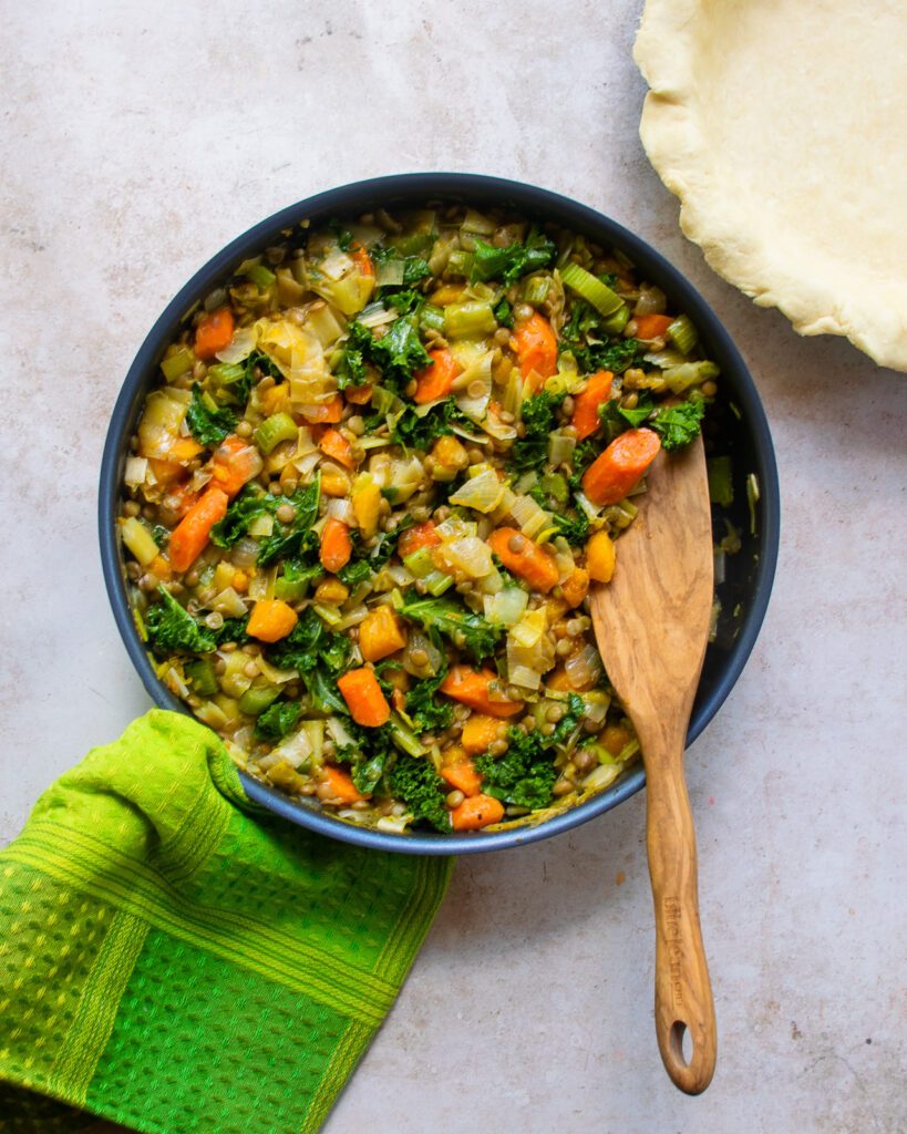 A blue skillet with vegetables and a wooden spoon, next to an empty pie plate filled with pastry.