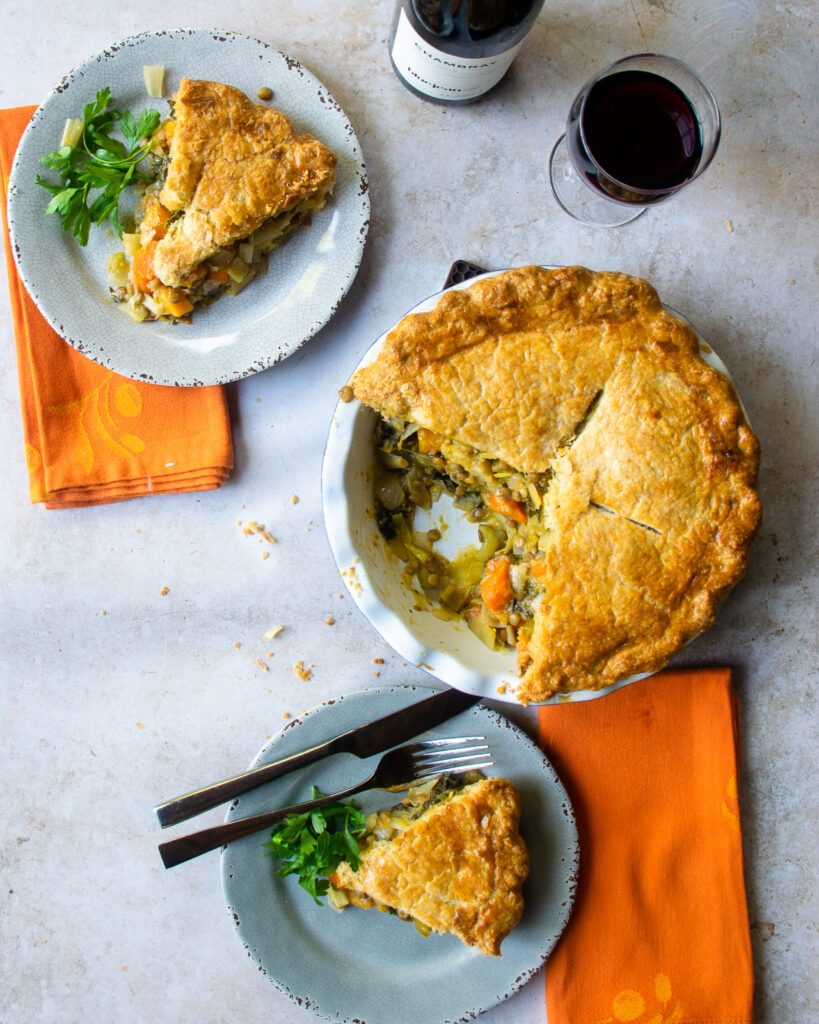 Looking down on two grey plates with a pie of vegetable pie, next to orange napkins and the rest of the whole pie with a bottle of wine.