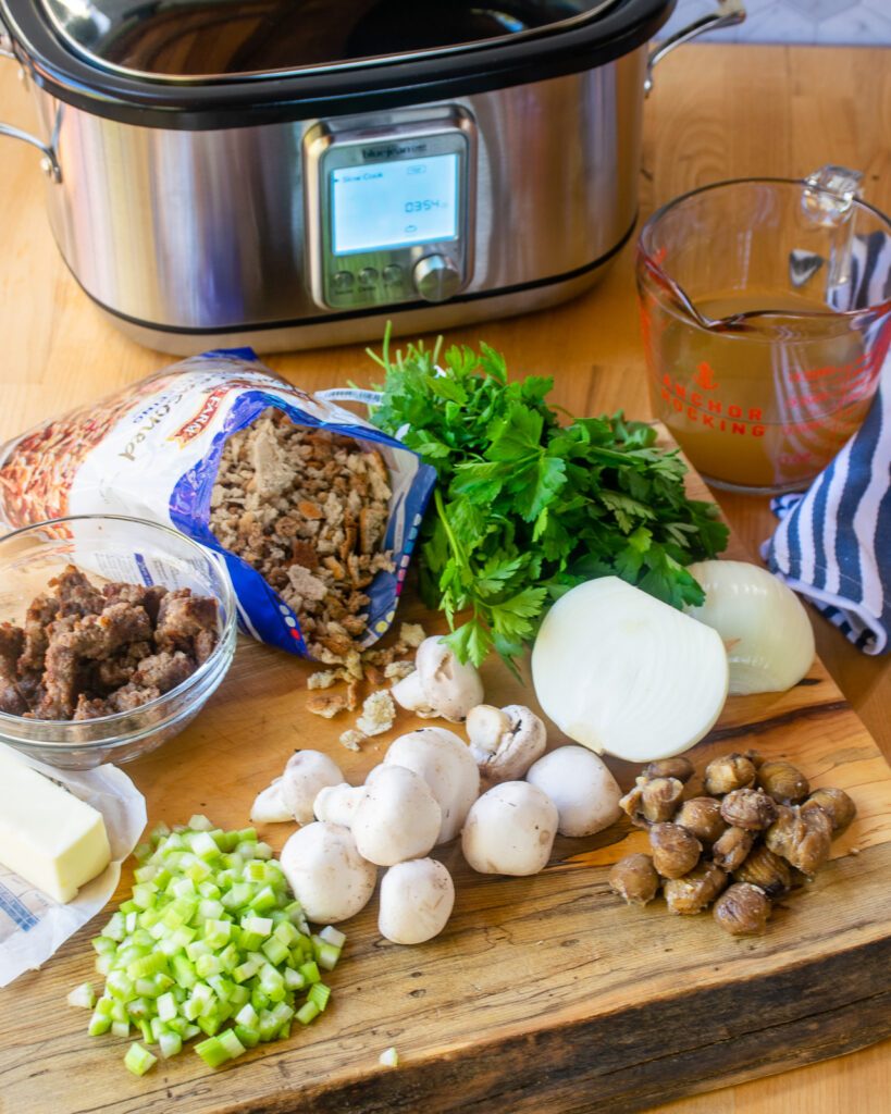 Ingredients on a cutting board in front of a slow cooker - stuffing bread, sausage, celery, mushrooms, chestnuts, onion, parsley and chicken stock.