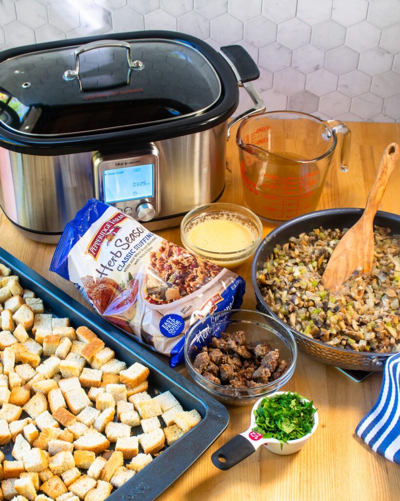 A slow cooker with ingredients in front of it - sauteed vegetables, beaten egg, chicken stock, bread cubes and parsley.