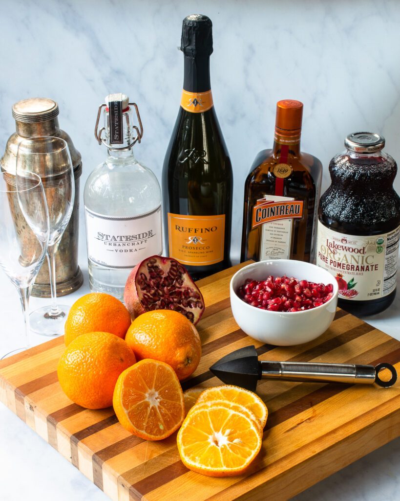Ingredients on a countertop - tangerines, pomegranate, pomegranate juice, cointreau, champagne, vodka and a cocktail shaker.