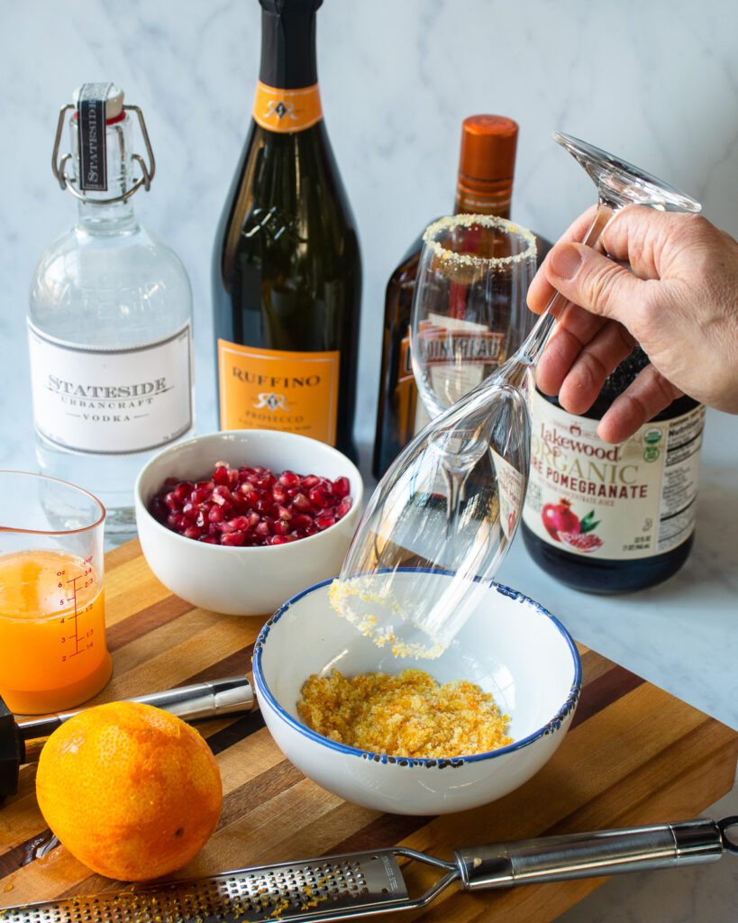 A hand dipping a champagne flute into sugared zest to coat the rim. Bottles in the background.