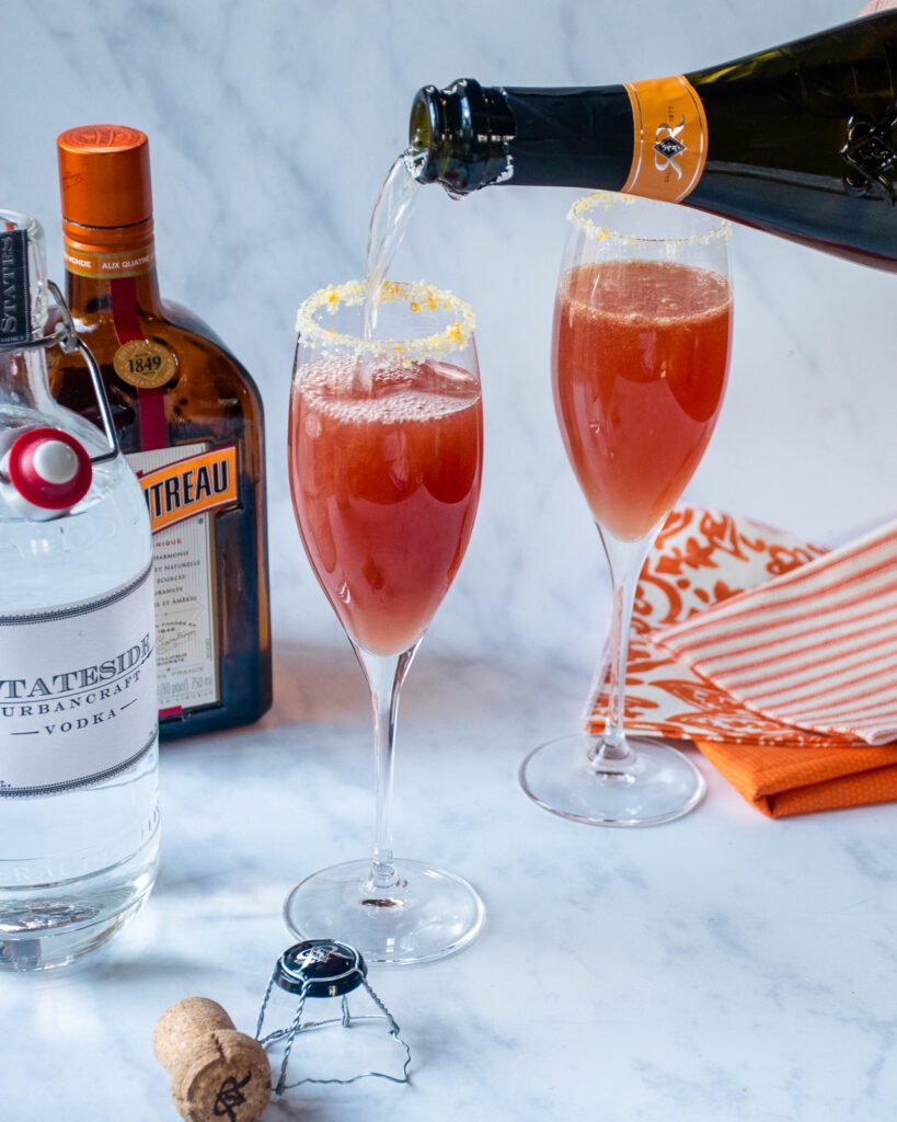 Topping a cocktail with champagne with bottles in the background.