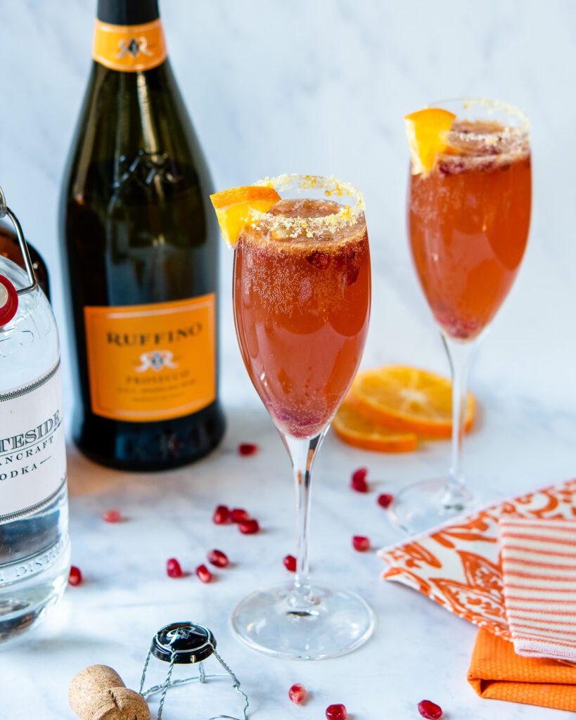 Two champagne flutes with a cocktail with a bottle of champagne and vodka in the background.
