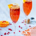 Two champagne flutes of pomegranate tangerine champagne cocktail on a marble counter with pomegranate seeds adn a few slices of tangerine scattered around.