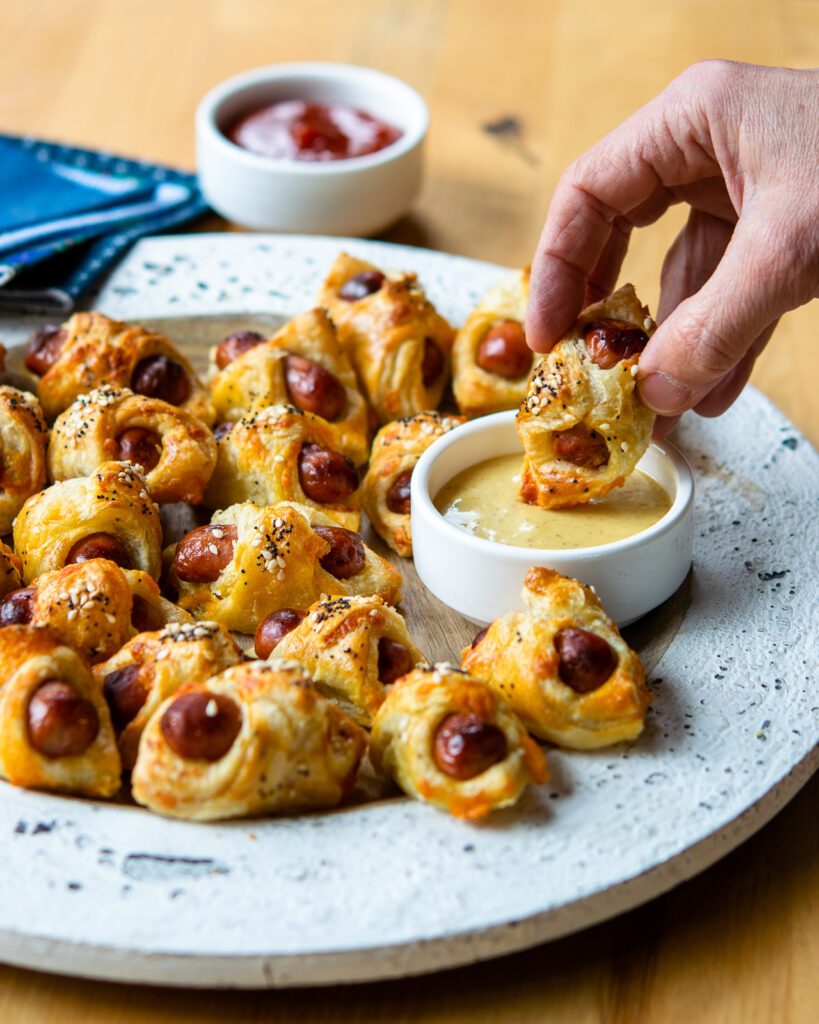 A hand dipping a pig in a blanket into honey mustard.
