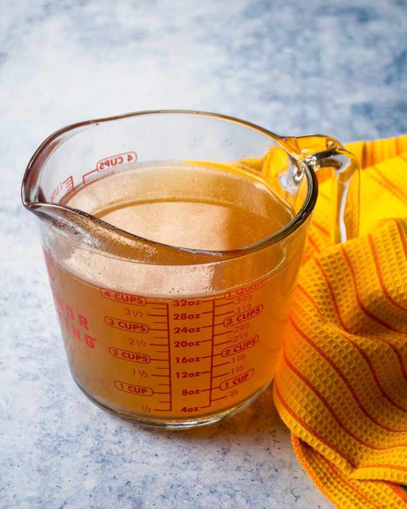 A measuring cup with chicken stock and a yellow and orange striped towel.