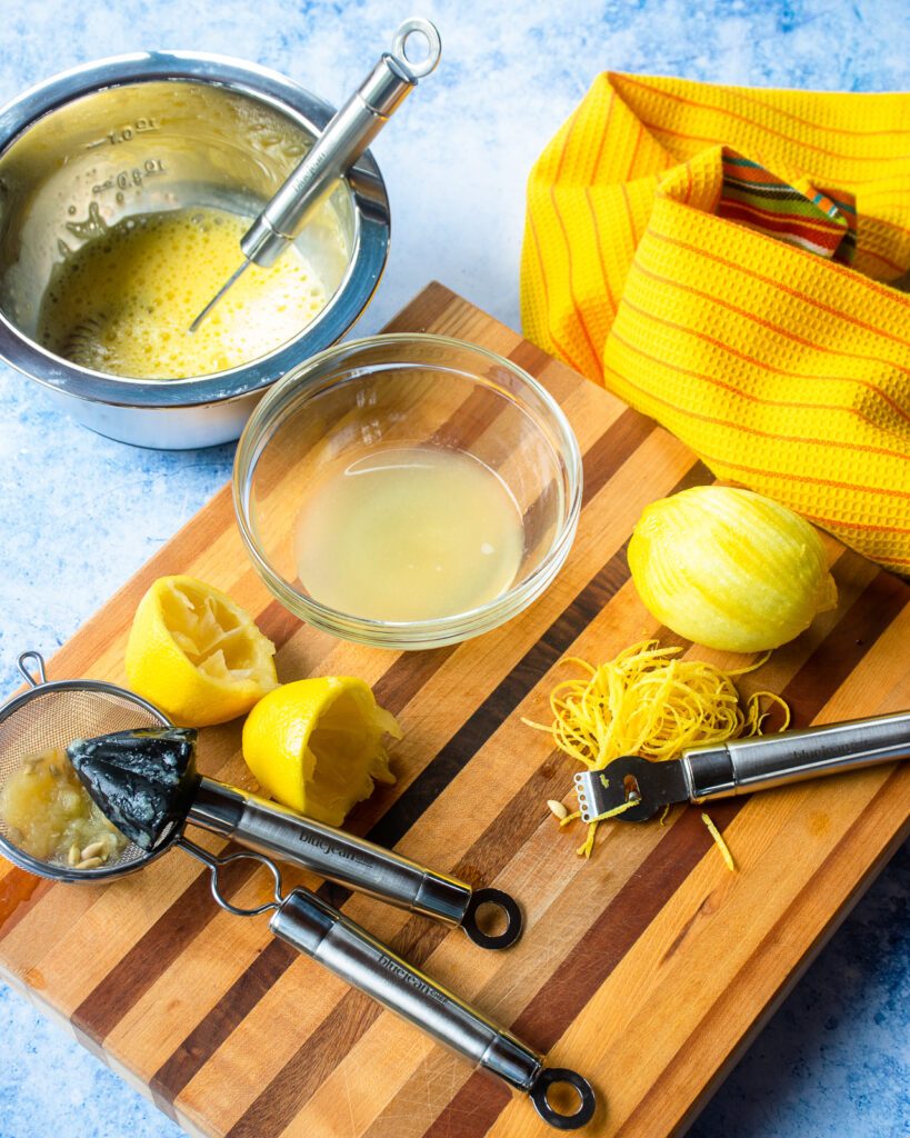 Several ingredients on a cutting board - lemon juice with reamer and strainer, zested lemon with a zester and a bowl of beaten eggs with a whisk.