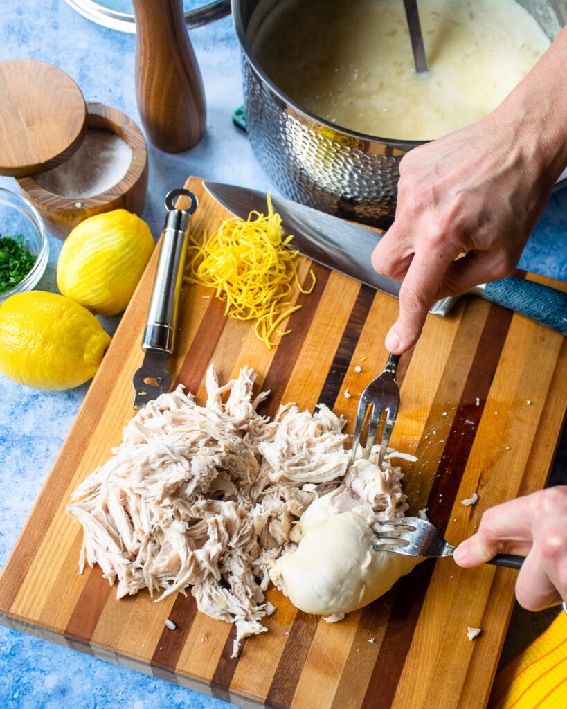 Hands shredding chicken breasts with two forks on a cutting board.