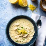 A blue bowl of avglolemono soup with a cut lemon, spoon, salt and pepper.