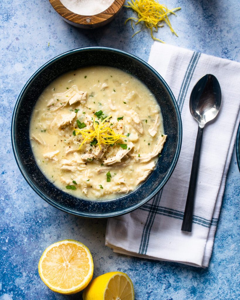 A blue bowl of avglolemono soup with a cut lemon, spoon, salt and pepper.