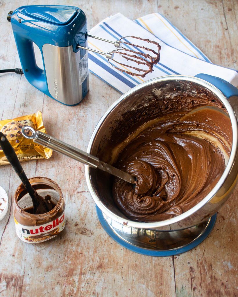 A pivoting mixing bowl with chocolate nutella frosting and a hand mixer near by.