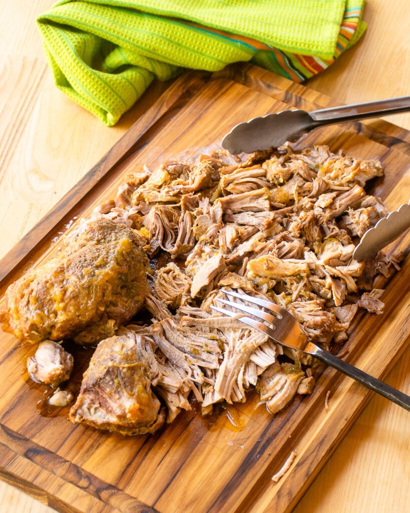 Pork butt being shredded on a wooden cutting board with a green towel in the background.