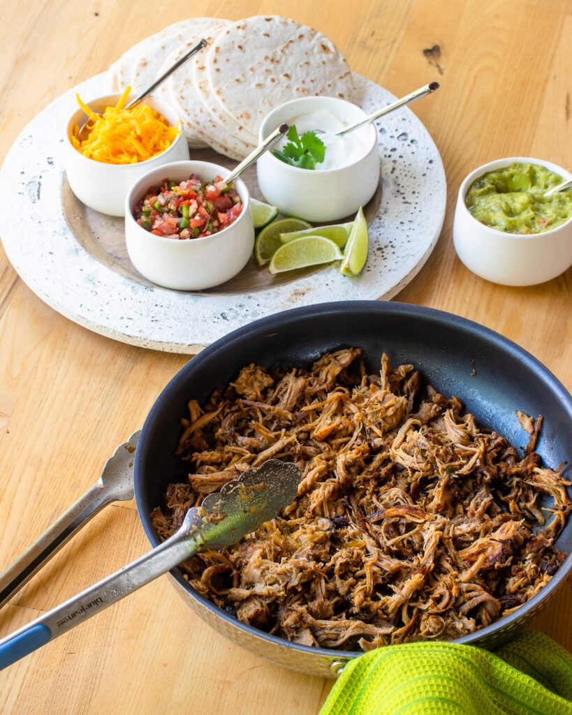 Pork carnitas in a skillet with taco fixings in the background.