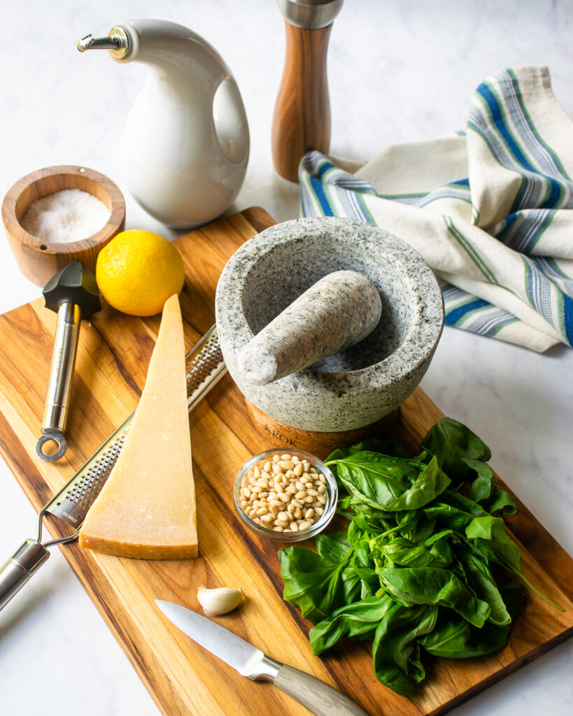 Ingredients for pesto on a board surrounding a stone mortar and pestle - basil, pinenuts, garlic, parmesan cheese, lemon, salt, pepper and olive oil.