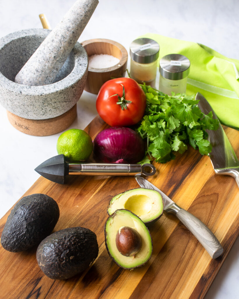 All the ingredients for guacamole on a cutting board - avocado, tomato, red onion, lime, cumin, cayenne pepper, salt - some knives, a reamer and a mortar and pestle.