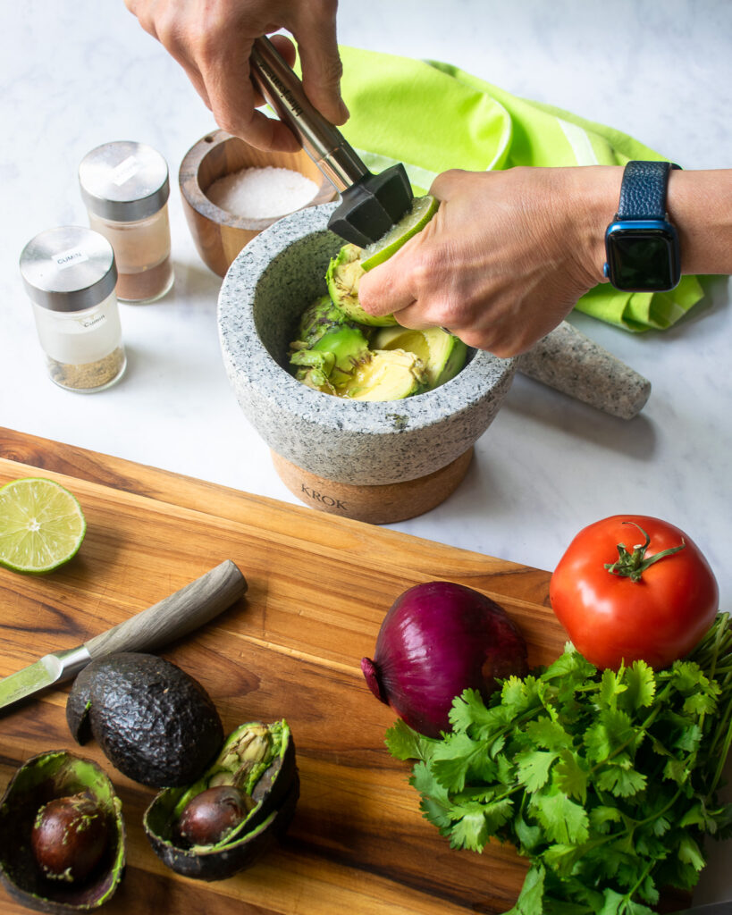 Hands using a reamer to squeeze lime juice into a mortar that has avocado in it. Other ingredients for guacamole nearby.