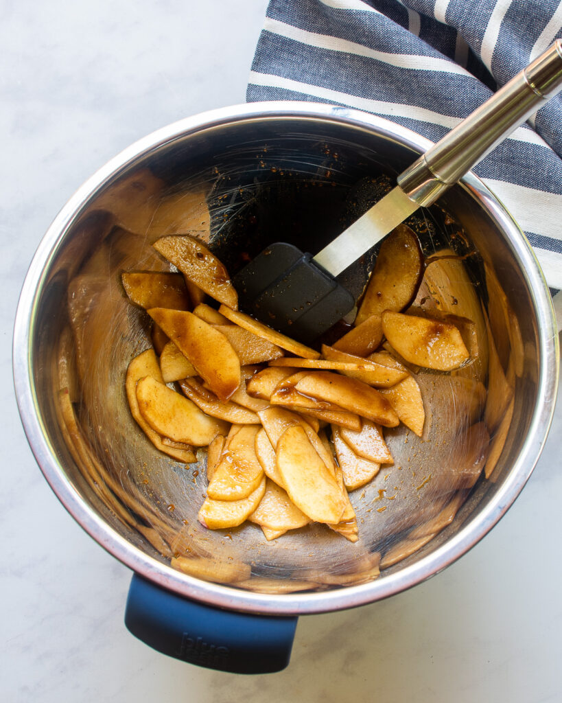 Looking down into a stainless steel bowl of apple slices tossed with brown sugar and spices with a spatula sticking out of the bowl and a blue and white striped towel nearby.