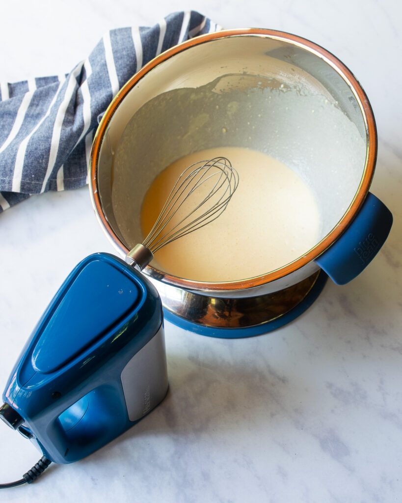 A stainless steel bowl with blue handle containing an egg custard, with a blue hand mixer with whisk attachment in front of it and a blue and white striped kitchen towel in the background.