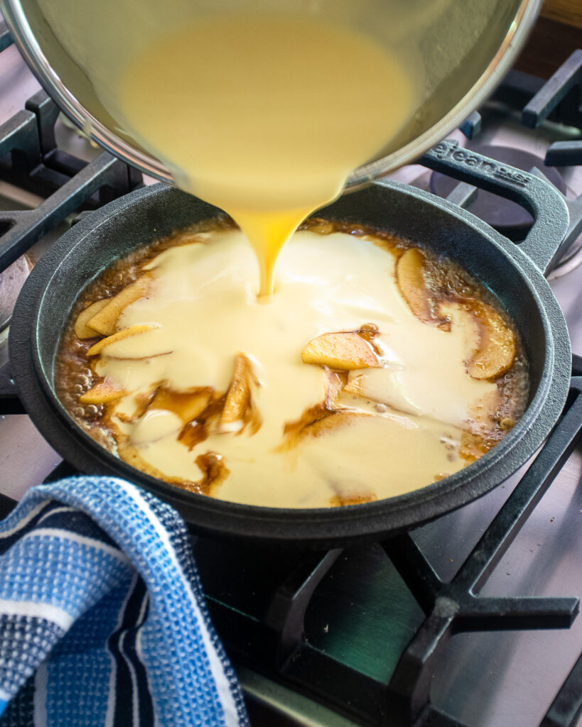A bowl pouring batter into a cast iron pan on a gas stovetop with sliced apples inside.