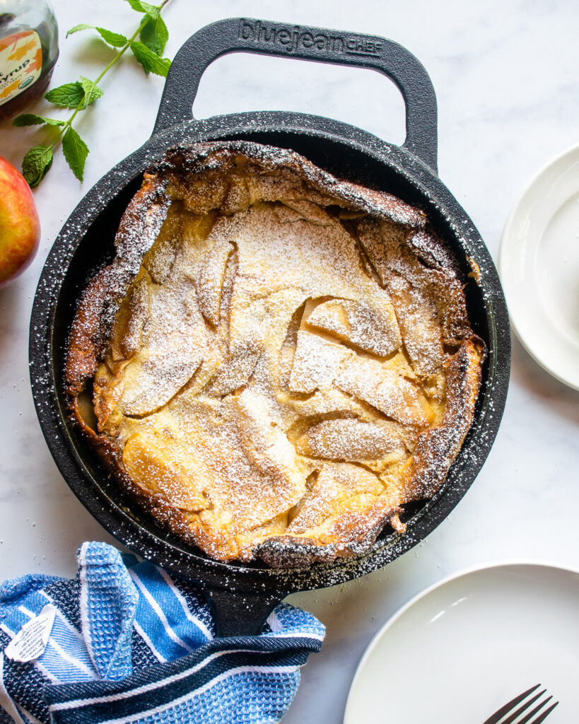 Looking straight down on an apple Dutch baby pancake in a cast iron pan with a blue kitchen towel wrapped around the handle, two white plates.