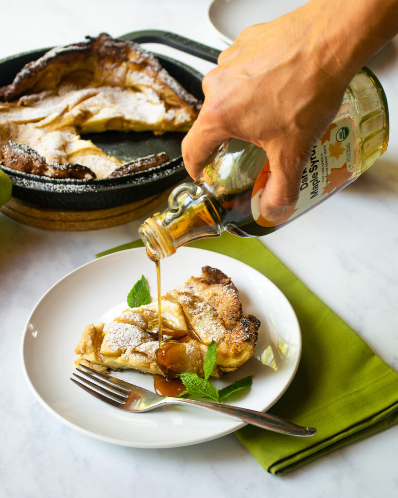 A hand pouring maple syrup on a slice of apple Dutch baby pancake on a white plate with a green napkin underneath and a cast iron skillet of more apple Dutch baby in the background.