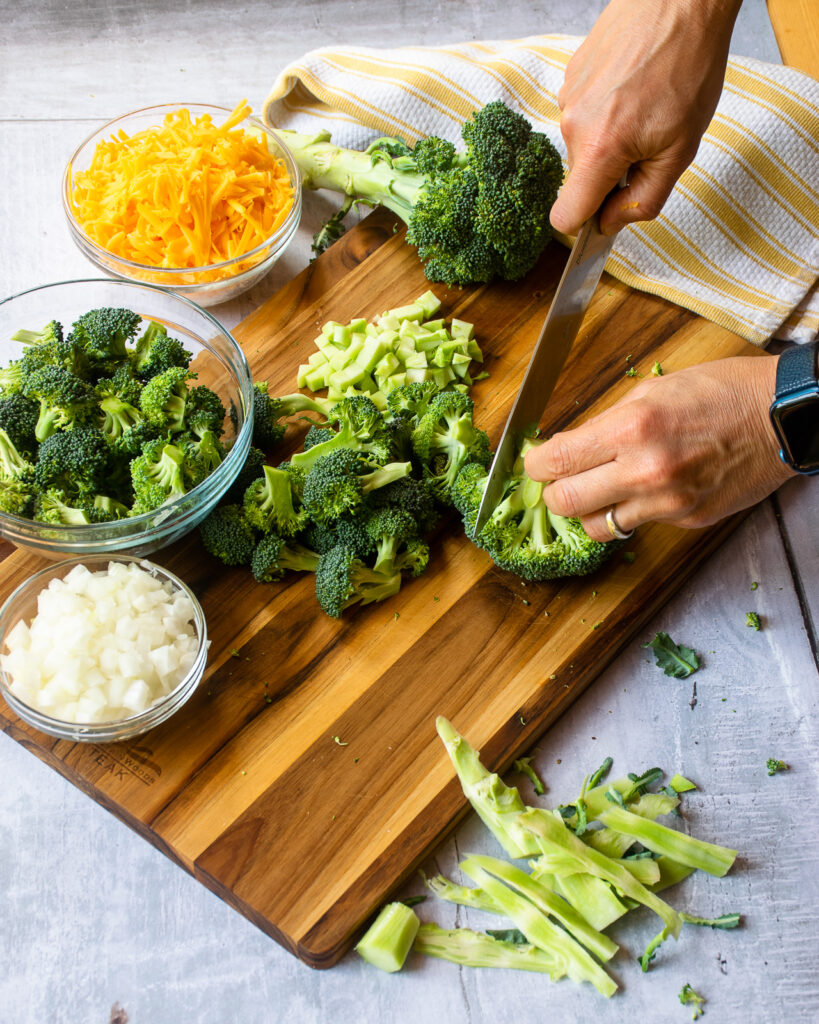 Hands cutting broccoli on a cutting board with a bowl of broccoli florets, a bowl of grated Cheddar, a bowl of diced onion and a yellow and white striped towel.