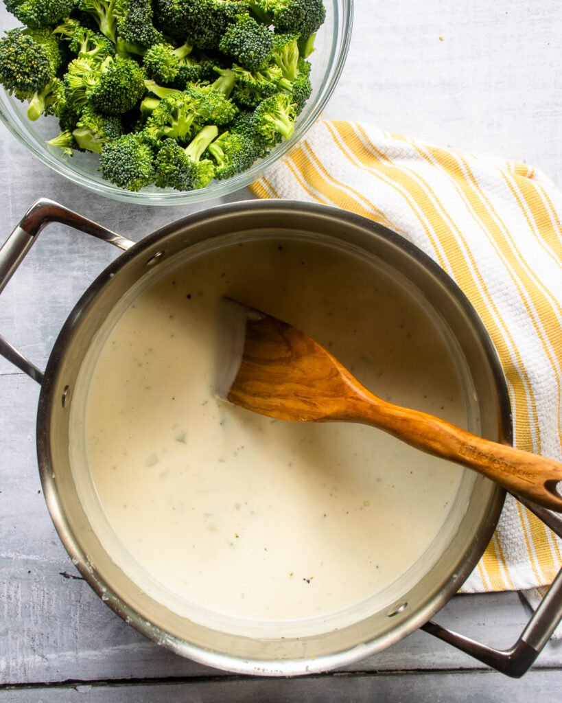 Looking down into a stockpot with a cream sauce and a wooden spoon, next to a bowl of broccoli florets and a yellow and white striped kitchen towel.