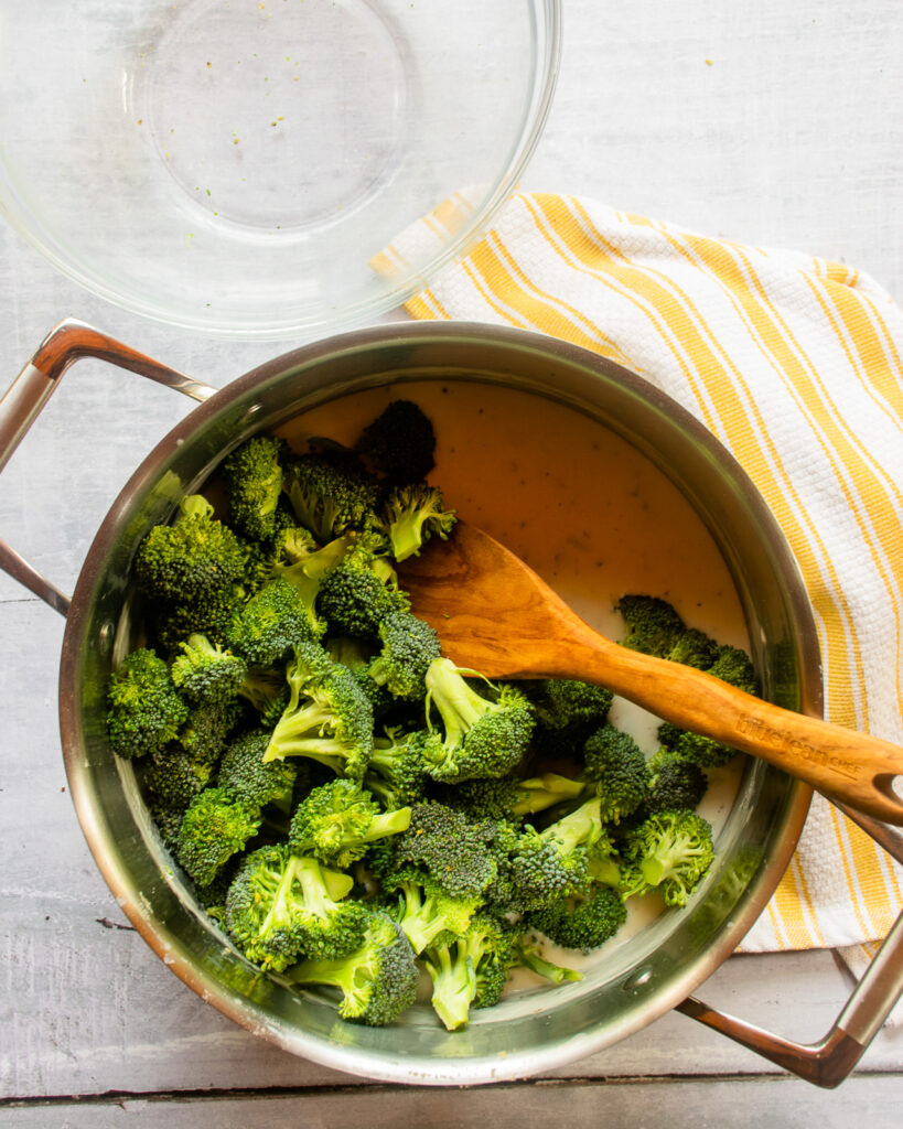 Looking down into a stockpot with broccoli florets and a white sauce and wooden spoon, next to a white and yellow kitchen towel.