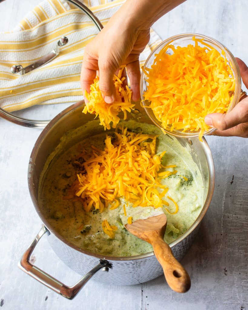 A hand adding Cheddar cheese to a stockpot of broccoli soup with a yellow and white striped kitchen towel and pot lid in the background.