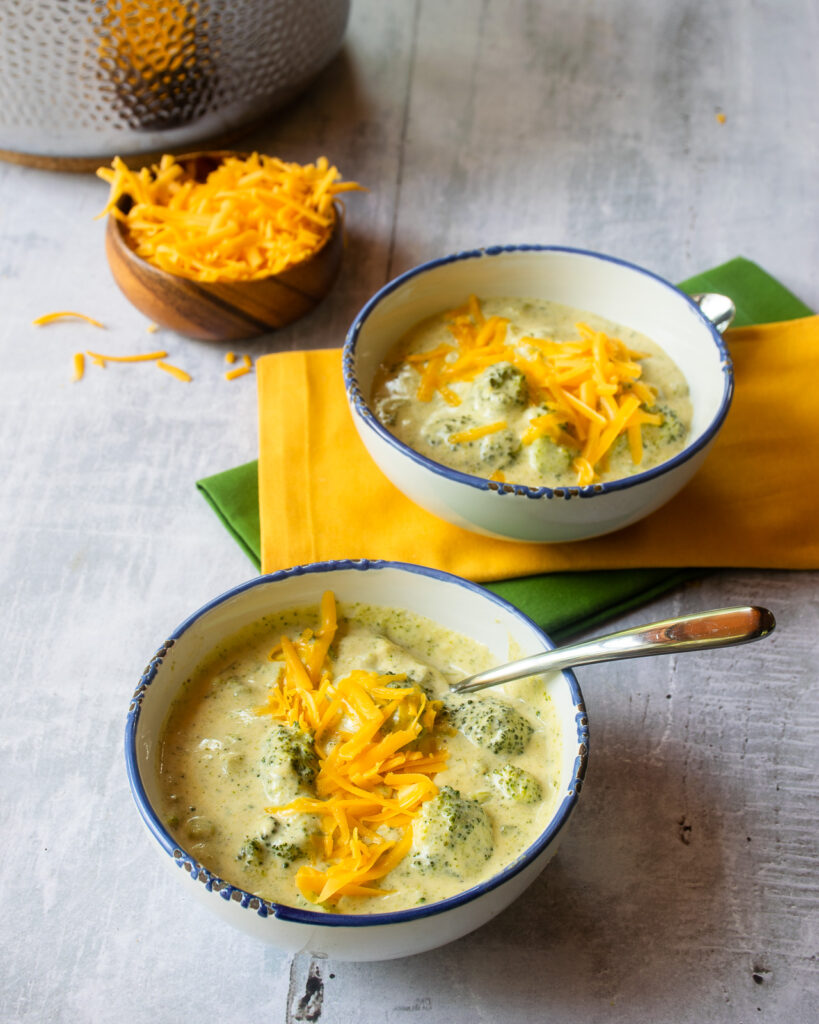 Two blue-rimmed white bowls of chunky broccoli Cheddar soup with yellow and green napkins, a small bowl of grated Cheddar cheese and a hammered stainless steel stockpot in the background.