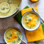 Looking down on two blue-rimmed white soup bowls with broccoli Cheddar soup, yellow and green napkins and a small bowl of Cheddar cheese.