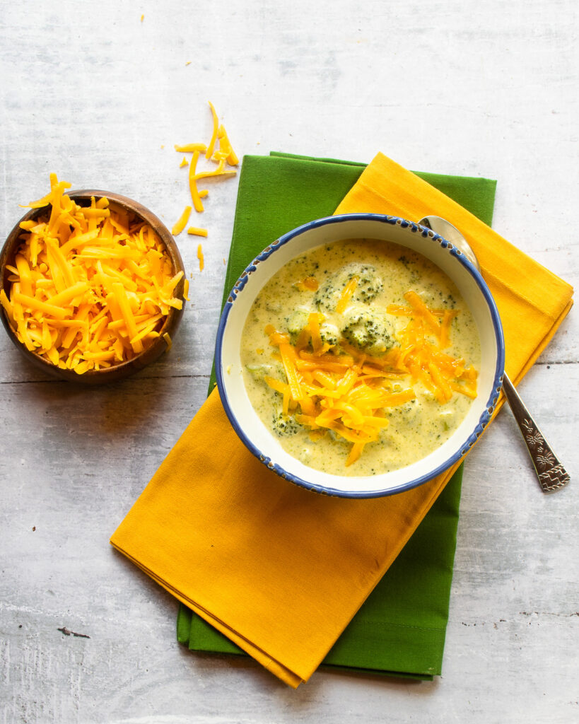 Looking down at one bowl of chunky broccoli Cheddar soup on yellow and green napkins and a small bowl of grated Cheddar cheese.